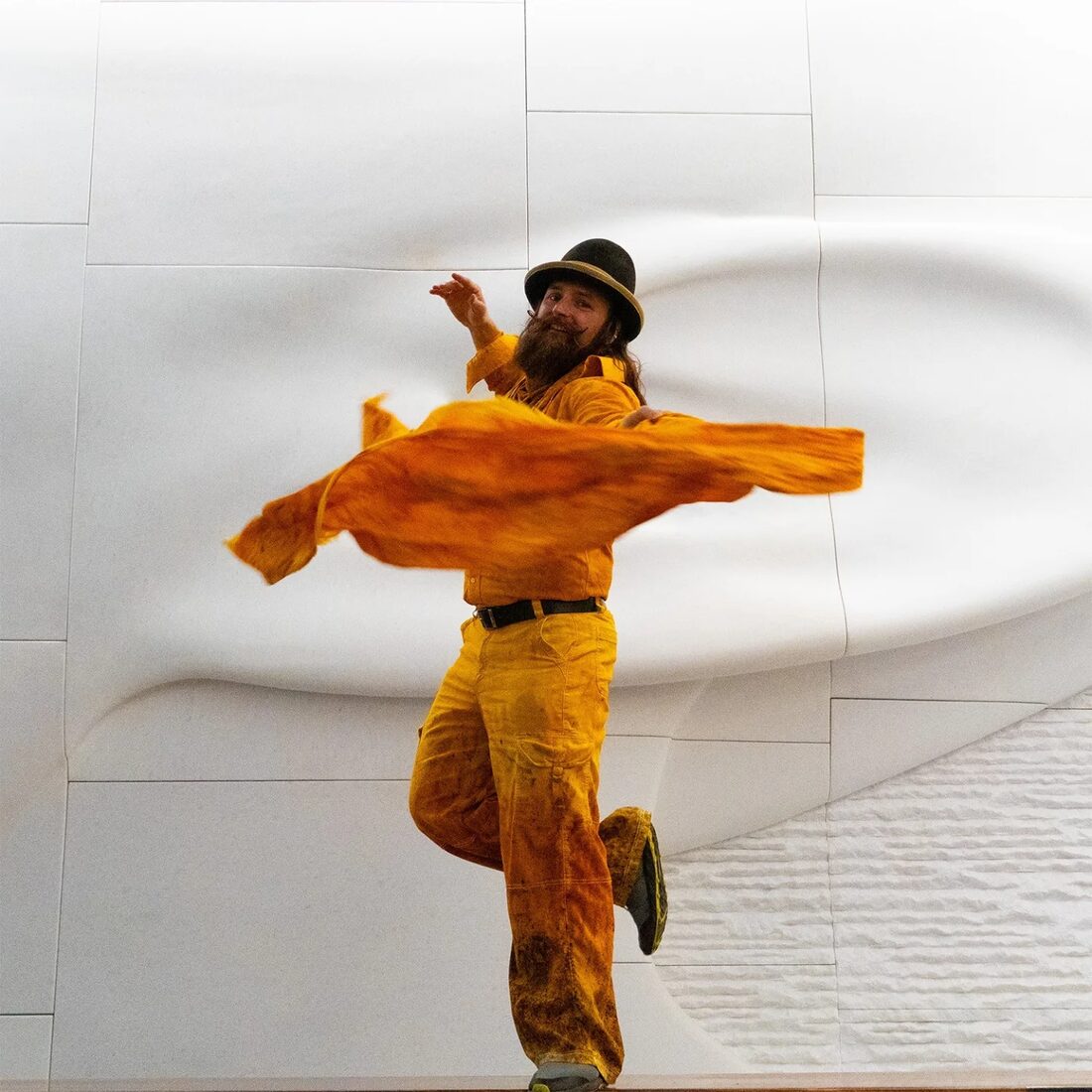 Scott Van Sice in a marigold suit and bowler hat, mid-spin with flowing fabric